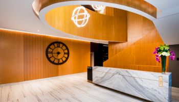Reception desk in a modern lobby with warm wood walls, neon globe light, a wall clock, and a vase of flowers.