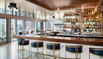 A modern bar with shelves full of liquor bottles, a countertop, and stools beside large city-facing windows.