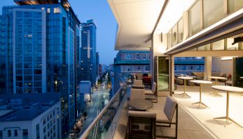 Rooftop terrace with outdoor seating overlooks a downtown street between tall glass office buildings at dusk.