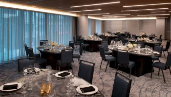 A modern event hall with banquet tables set for dinner&mdash;place settings, glasses, candles, and floral centerpieces in a sleek room.