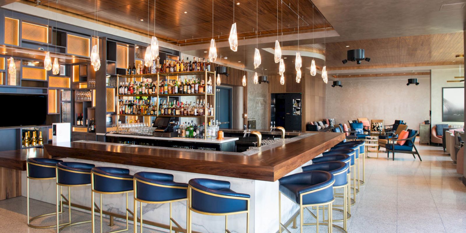A modern bar lounge with a wooden counter, backlit liquor shelves, pendant lights, stools, and a seating area.