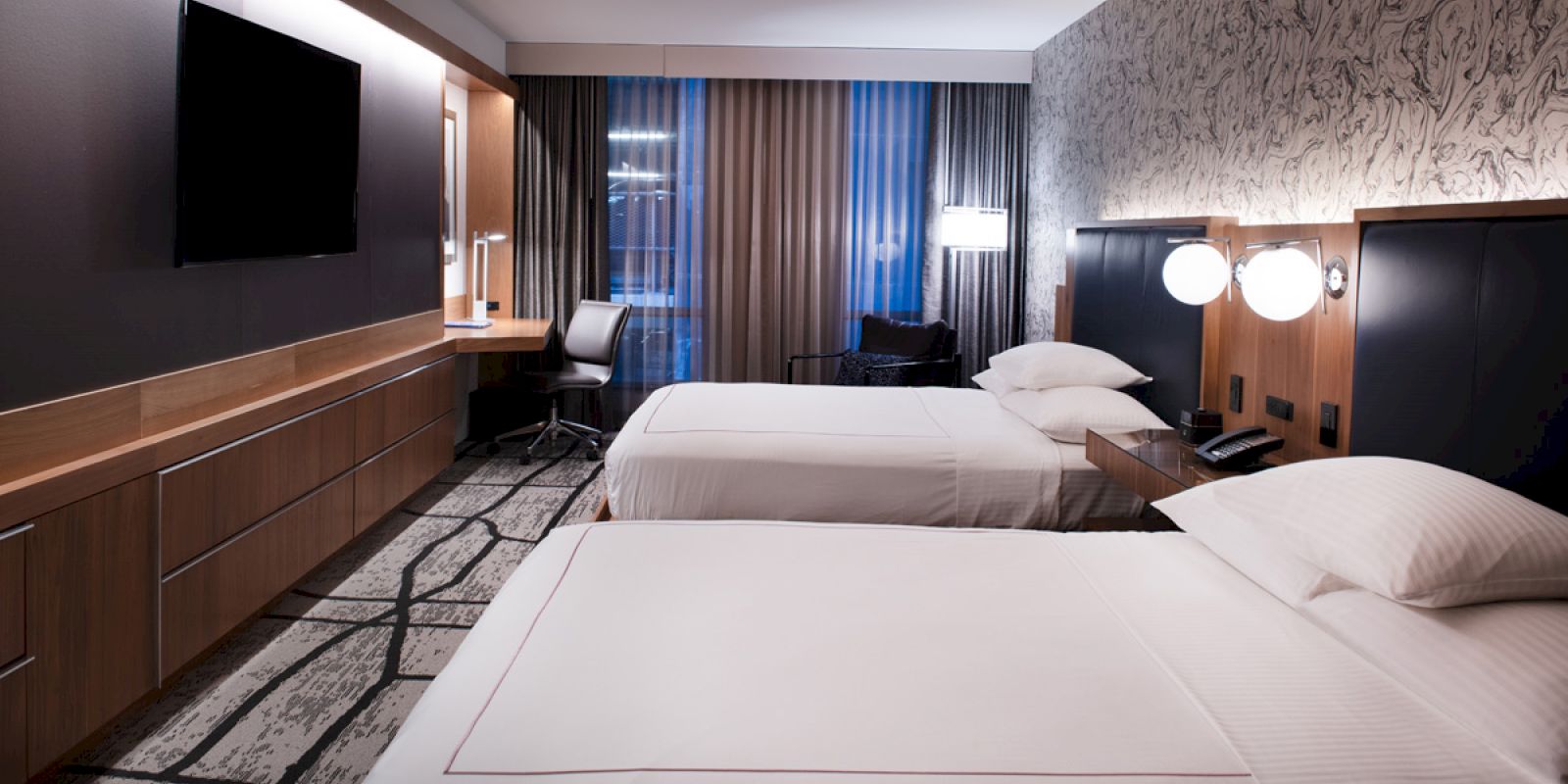 A modern hotel room with two neatly made beds, dark wood panels, and a desk, lit by bedside lamps.