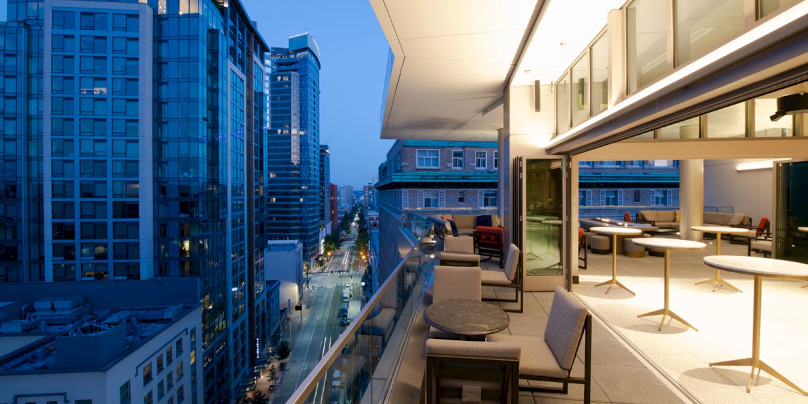 A modern high-rise balcony overlooks a city street at dusk, with outdoor lounge chairs and a sleek rooftop terrace.