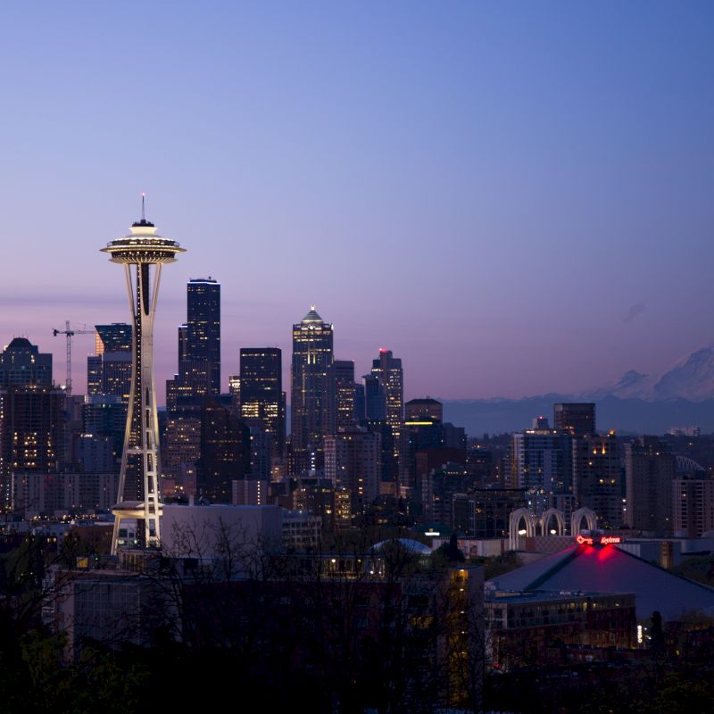 Seattle&rsquo;s skyline at dusk, with the Space Needle glowing above downtown buildings and distant mountains under a purple sky.