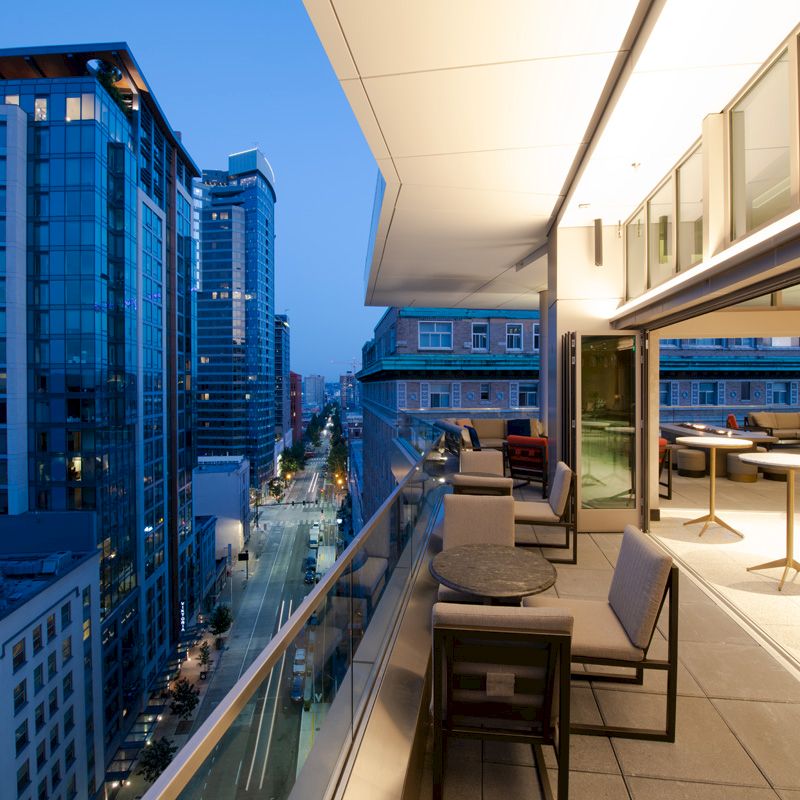 A modern high-rise rooftop terrace overlooks a city street and skyscrapers, with outdoor seating and large windows at dusk.