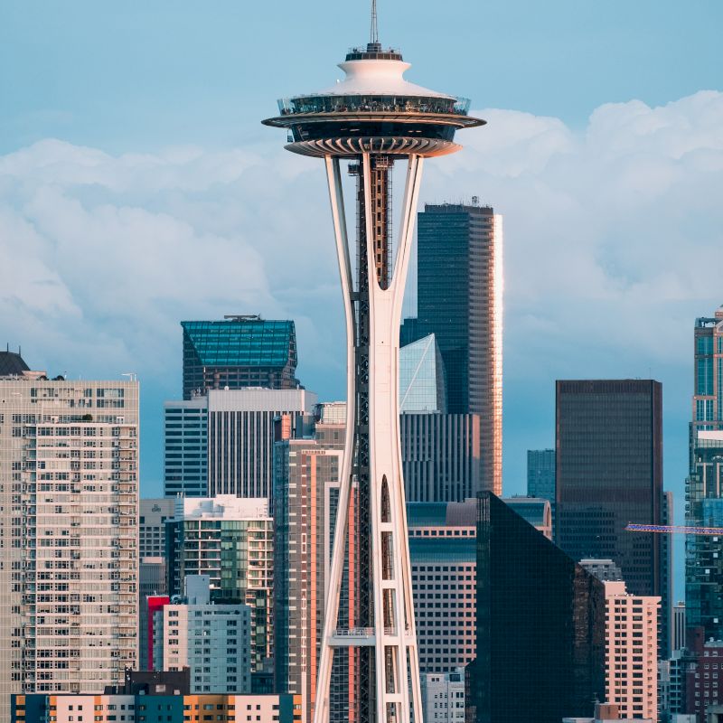 A view of Seattle&rsquo;s Space Needle rising above city buildings under a blue sky with clouds.