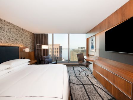 Modern hotel room with a bed, desk chair, built-in TV, and large windows overlooking city buildings.