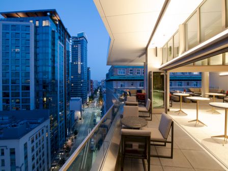 A modern high-rise rooftop terrace with chairs and a view down city streets between buildings, at blue-hour dusk.