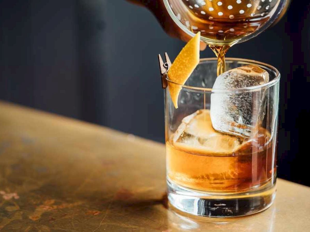 A golden cocktail is being poured into a glass over ice, with an orange garnish triangle on top, and the drink is amber.