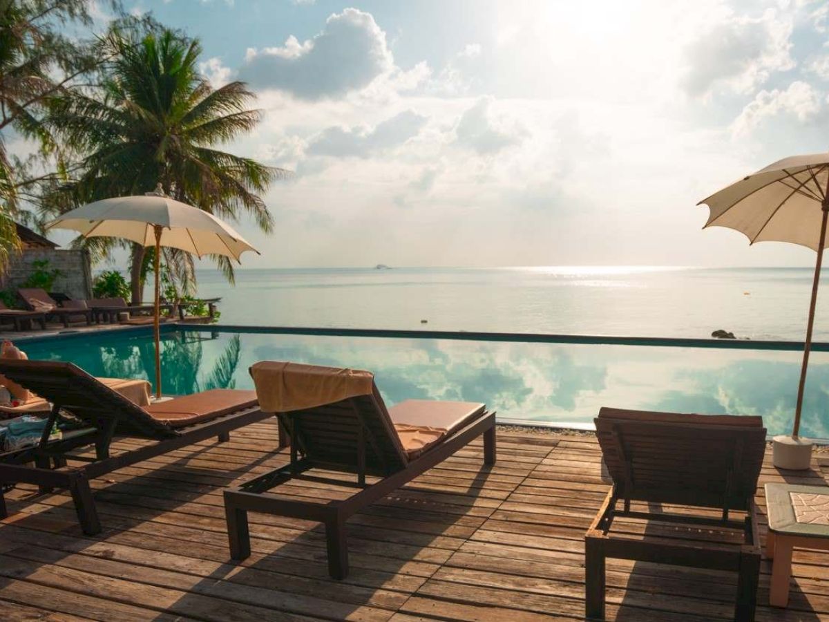 Tropical resort pool deck with sun loungers and two umbrellas overlooking the ocean under a bright, cloudy sky.