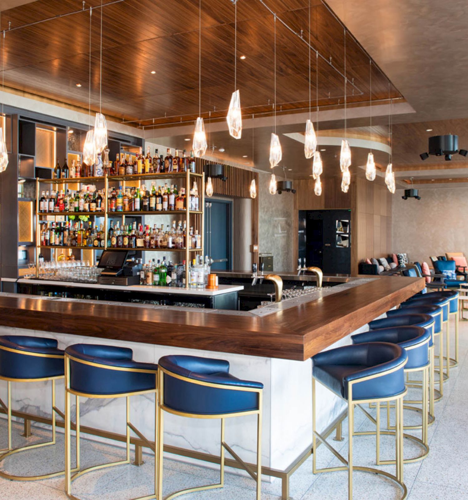A modern hotel bar with a wooden counter, hanging pendant lights, a full bottle shelf, and blue stools in a lounge.