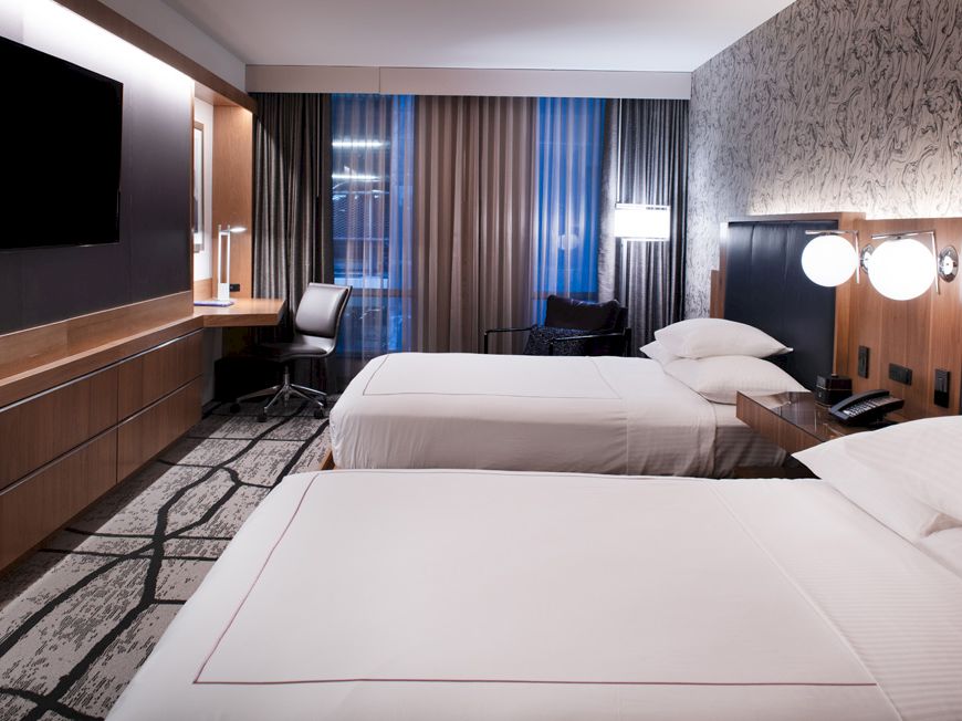 A modern hotel room with two beds, wood paneling, a TV, a desk chair, and blue curtains.