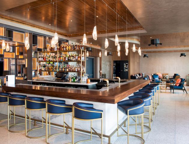 A modern hotel bar with a wooden counter, hanging lights, stocked shelves, and blue leather stools.