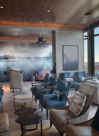 Modern lounge with comfy chairs, a skyline mural, and floor-to-ceiling windows overlooking a river at sunset.