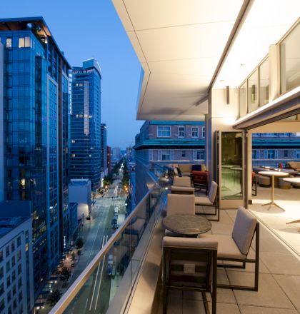 Modern high-rise terrace with outdoor seating overlooks a city street at dusk, with tall blue glass buildings in view.