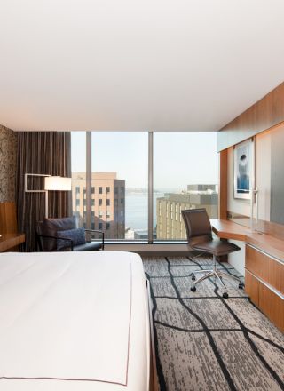 Modern hotel room with a large bed, desk chair, two wall TVs, and a city-view window.