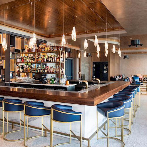 A modern hotel bar with a long wooden counter, bottle shelves, hanging pendant lights, and blue high stools in a lounge.