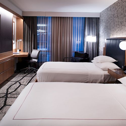 Modern hotel room with twin beds, curtained windows, and a wall-mounted TV.