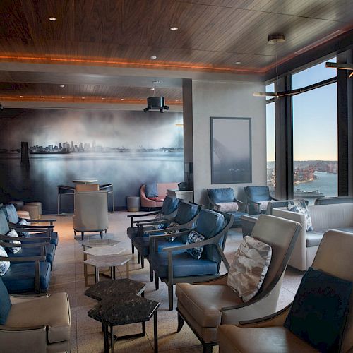 Modern lounge with leather chairs and tables, a skyline mural, and large windows overlooking a river at dusk.