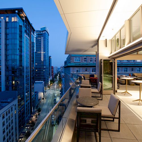 Modern rooftop terrace with outdoor seating overlooking a city skyline of tall buildings under a twilight-blue sky.