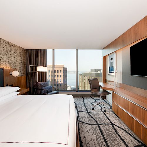 A modern hotel bedroom with a large bed, desk chair, wall TVs, wood paneling, and big city-view windows.
