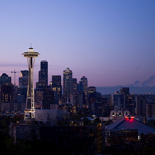Seattle skyline at dusk with the Space Needle towering above office buildings and distant mountains under a purple sky.