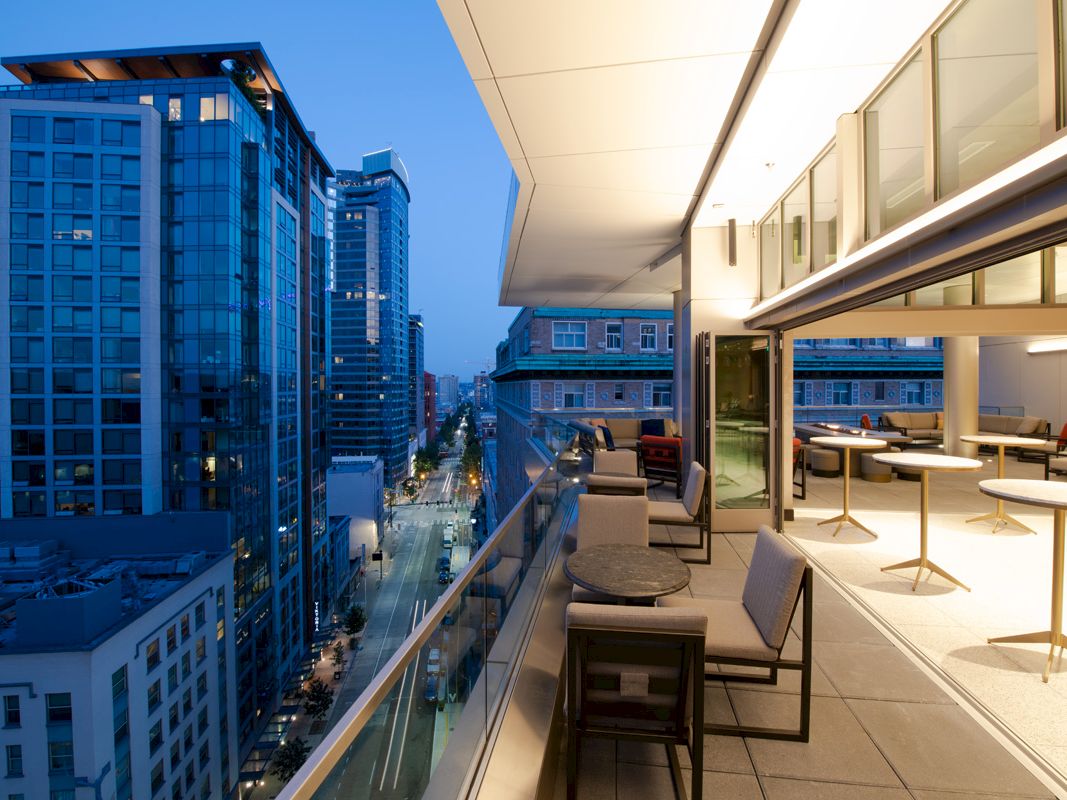A modern high-rise rooftop terrace with lounge seating overlooks a bright downtown street and skyscrapers.