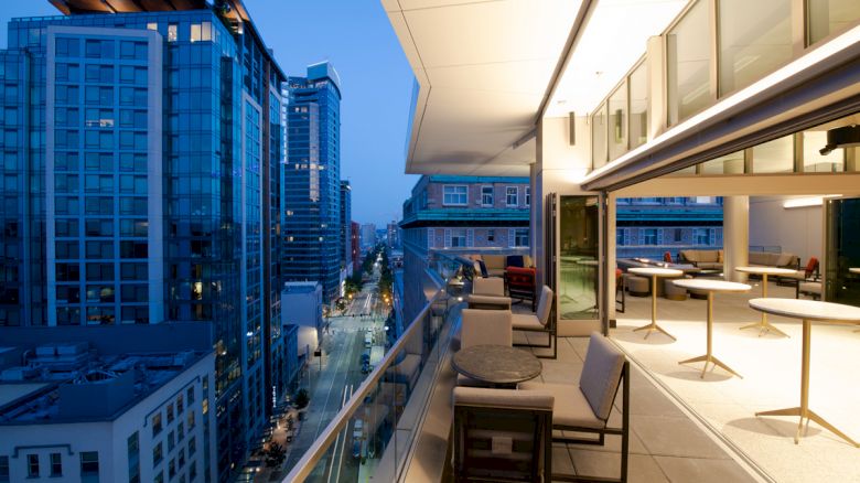 A modern high-rise rooftop terrace with lounge seating overlooks a bright downtown street and skyscrapers.