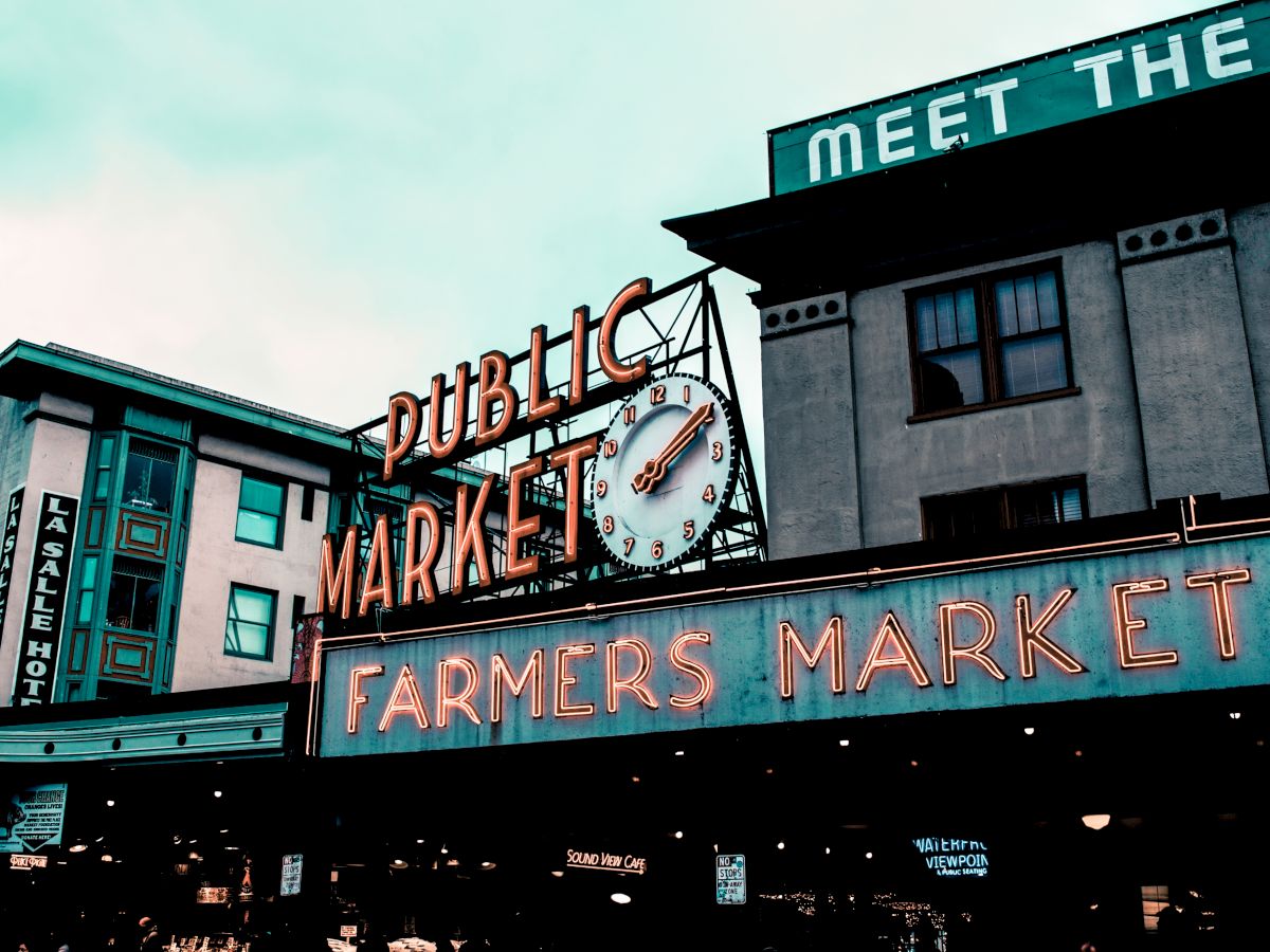 Neon signs on a storefront read &ldquo;Public Market&rdquo; and &ldquo;Farmers Market,&rdquo; with a clock over an urban building.