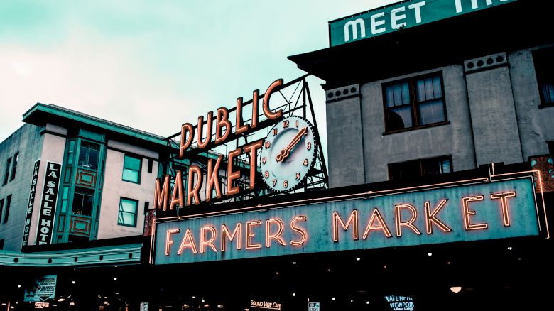 Neon signs on a storefront read &ldquo;Public Market&rdquo; and &ldquo;Farmers Market,&rdquo; with a clock over an urban building.