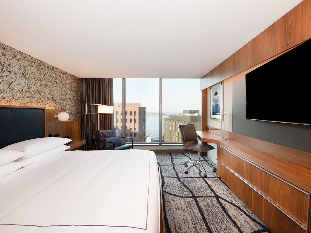 Modern hotel room: big bed, desk chair, wood TV wall, and floor-to-ceiling window with city view.