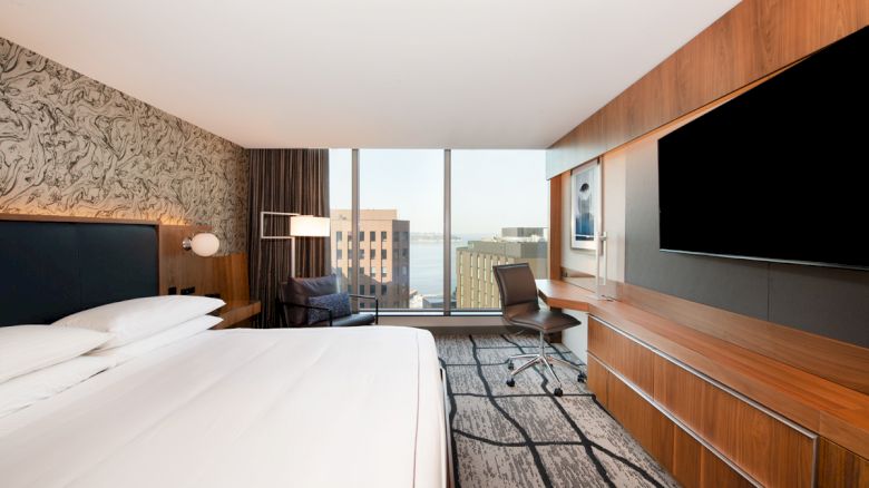 Modern hotel room: big bed, desk chair, wood TV wall, and floor-to-ceiling window with city view.