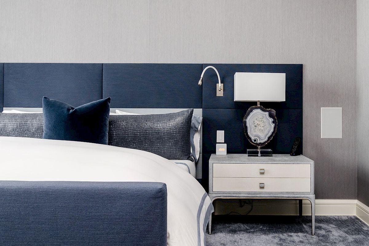 A modern blue bedroom with a made bed, decorative pillows, and a nightstand holding a lamp, clock display, and remote.