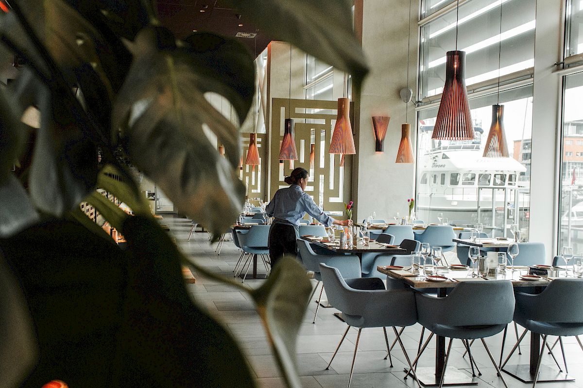 A modern restaurant dining area with neatly set tables, pendant cone lights, and a waiter serving near large windows and a bus outside.