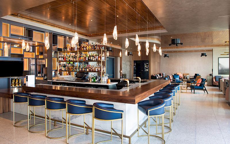 Modern hotel bar with a stocked liquor shelf, central wooden counter, blue stools, pendant lights, and lounge seating.