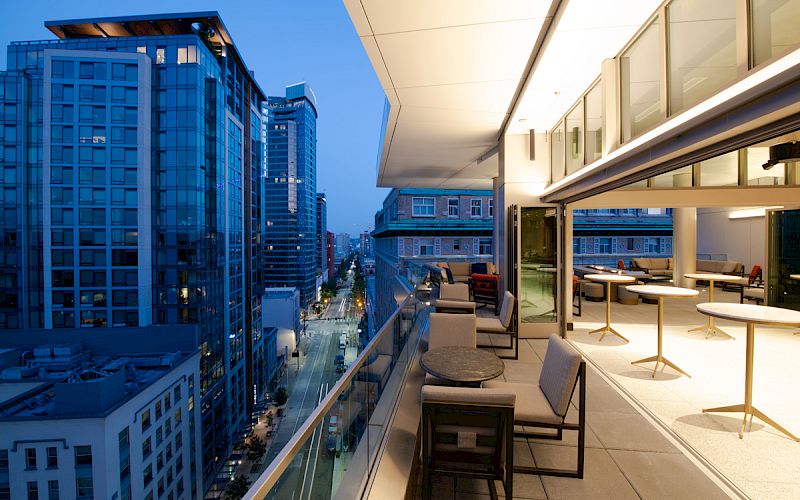 A modern high-rise rooftop terrace with lounge chairs overlooking a city street between tall buildings.