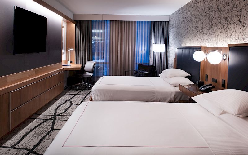 A modern hotel room with two white beds, black headboards, warm lighting, and floor-to-ceiling curtains.