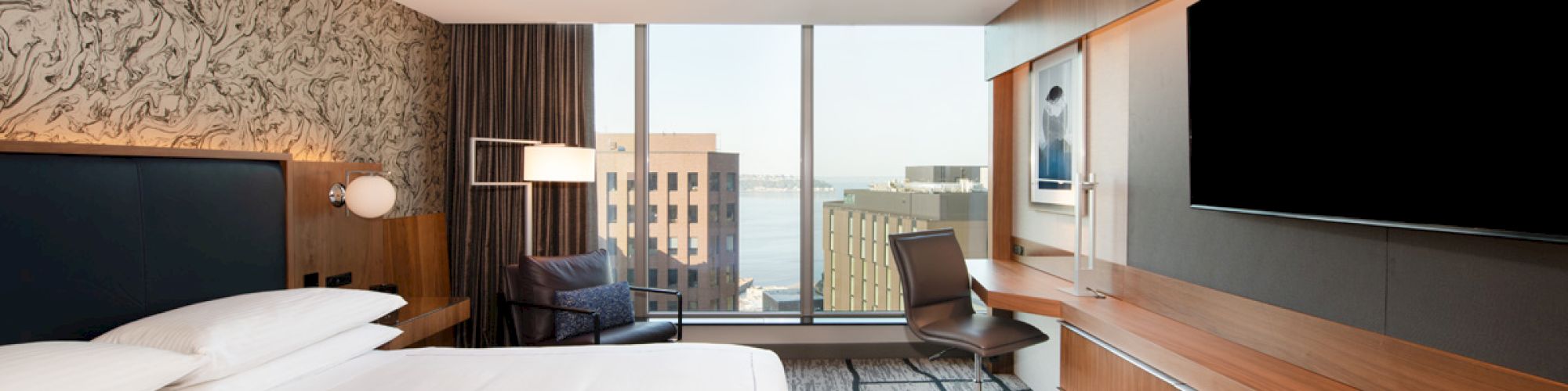 A modern hotel room with a large bed, desk chair, two TVs, and big windows overlooking city buildings.