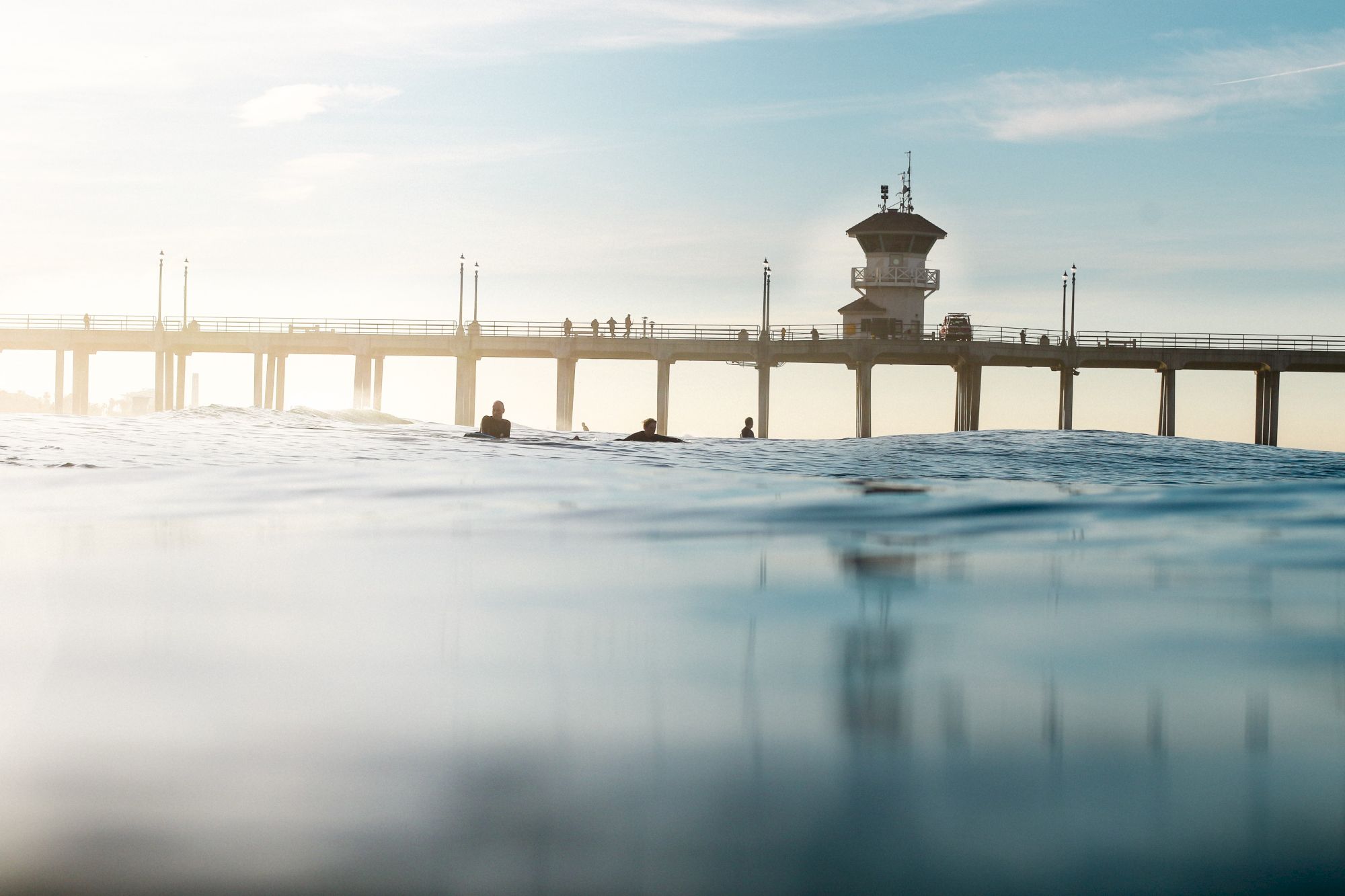A long pier and bridge over calm water with several swimmers near a lifeguard tower at sunset.